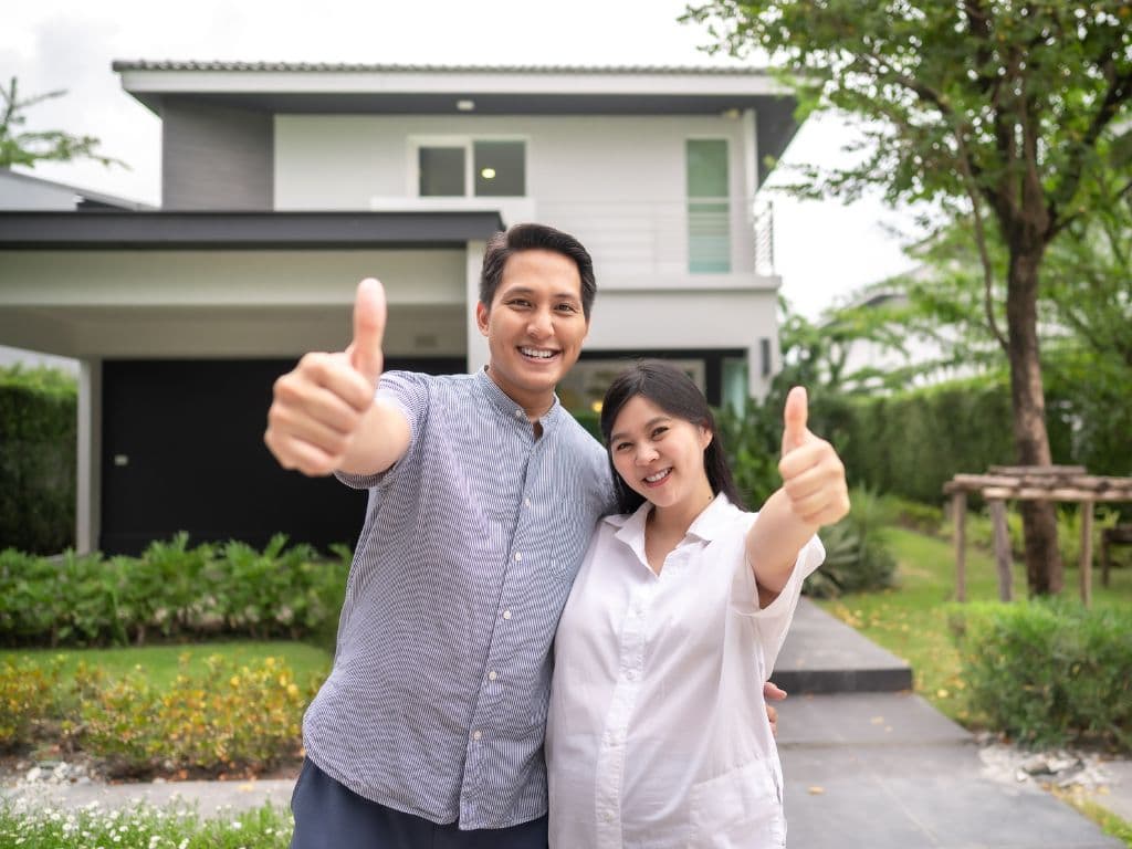 An Asian couple with a pregnant woman and their thumbs up in front of a house.
