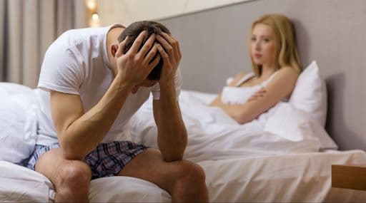 A man with his hands on his head as his girlfriend looks disappointed in bed
