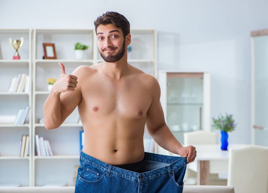 A shirtless man holding up jeans which no longer fit him because he is thinner while giving a thumbs up