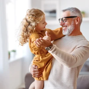 A loving grandfather with glasses and a white beard joyfully lifts his granddaughter in a mustard-yellow dress, sharing a warm moment in a cozy living room, reflecting the vitality supported by Vita Bella’s Metformin treatments.