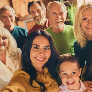 A multi-generational group of people smiling and taking a selfie in a cozy indoor setting with a brick wall and wooden background, representing family wellness.