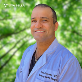 Dr. Irshad Shakir, Head Collaborating Physician at Vita Bella, wearing a white lab coat with his name and title as an Orthopedic Surgeon, against a green background